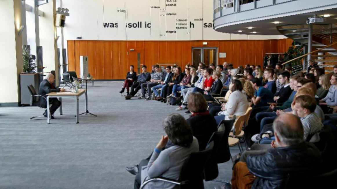 Zeitzeuge Sally Perel sitzt an einem Rednerpult vor einigen Menschen, welche ihm ebenfalls sitzend zuhören.