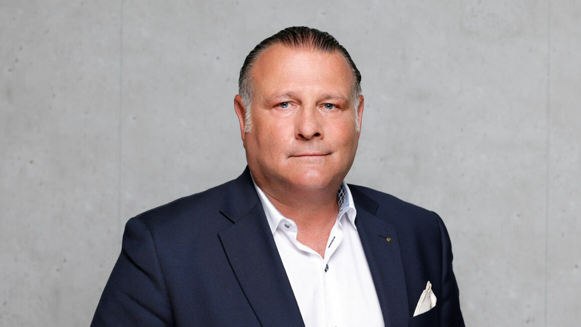 Das Bild zeigt Dr. Arno Homburg vor einer grauen Beton-Wand. Er trägt ein Anzug und ein Hemd.