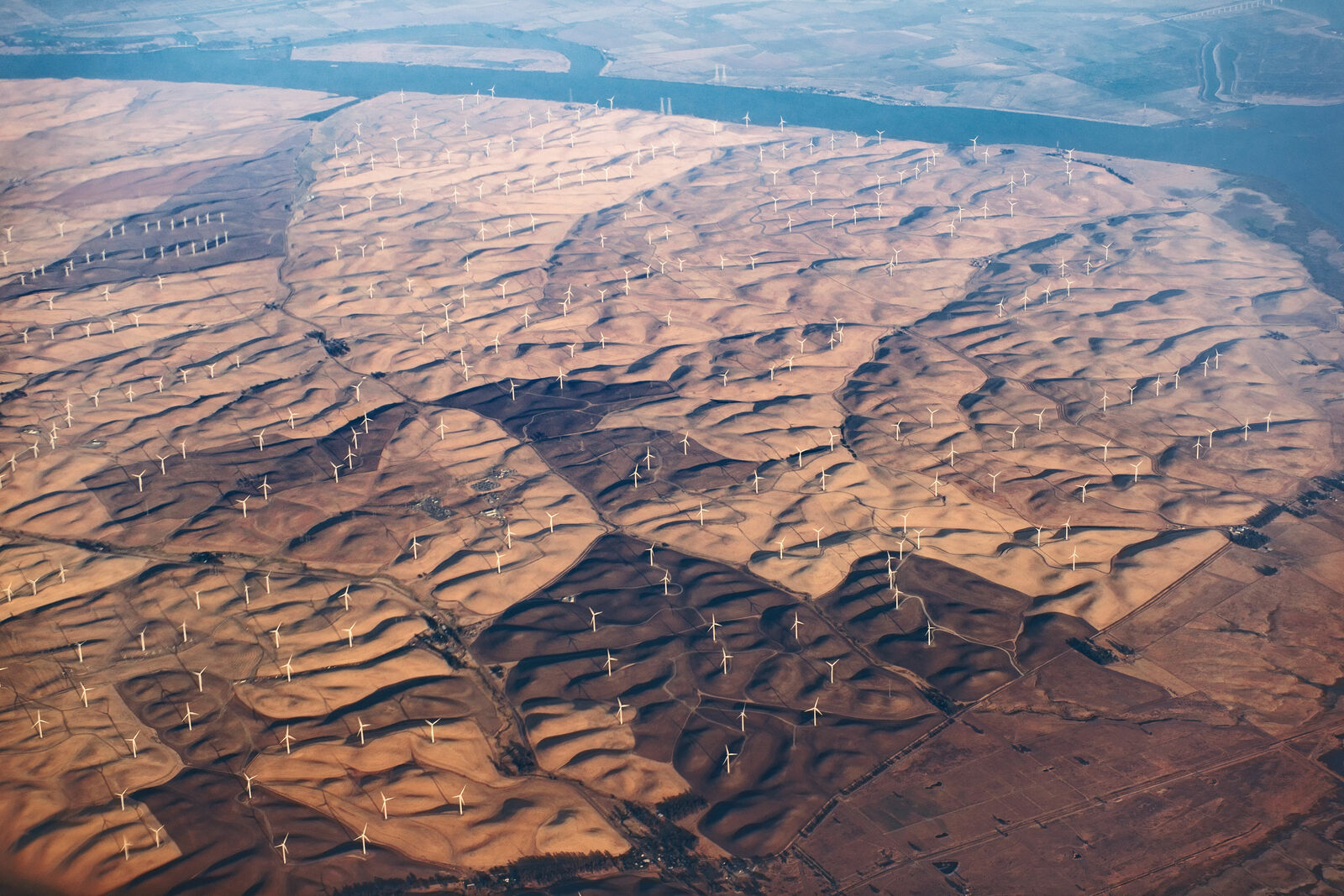 Aerial view of a vast wind farm in a hilly, arid landscape. Numerous white wind turbines are scattered across the sandy-colored hills. In the background, a large river flows.