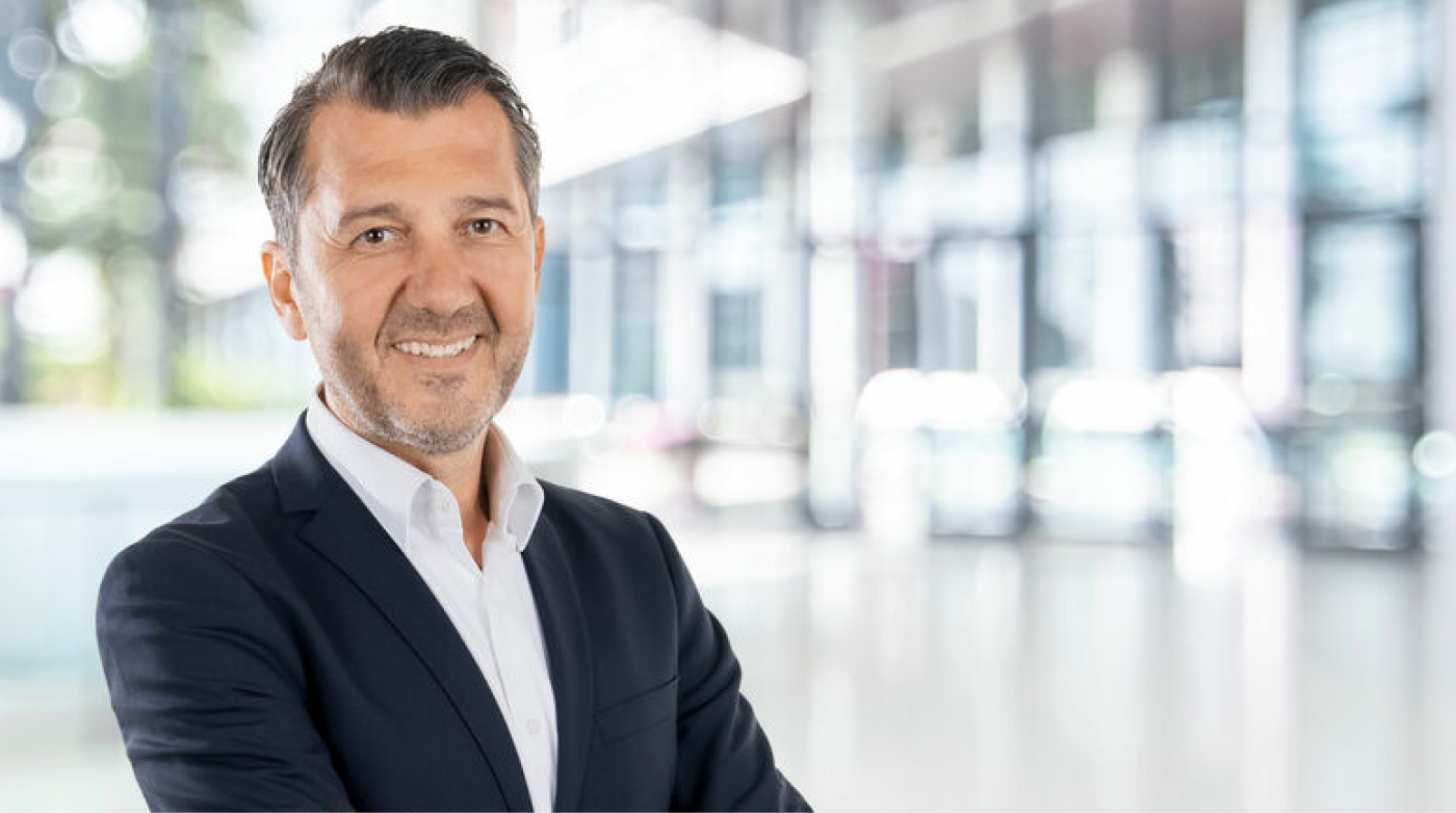 Ein Mann mittleren Alters mit dunklen Haaren und kurzem Bart steht in einem modernen Bürogebäude. Er trägt einen dunkelblauen Anzug mit einem weißen Hemd und lächelt freundlich in die Kamera. Der Hintergrund ist hell und verschwommen, mit großen Glasfenstern, die natürliches Licht hereinlassen.