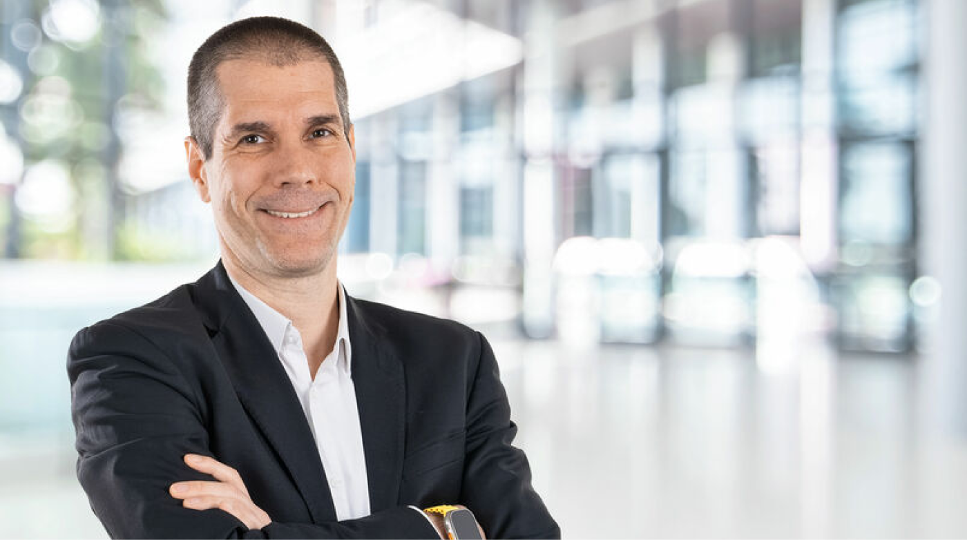 Porträt eines lächelnden Mannes im Business-Outfit mit verschränkten Armen vor einem modernen, lichtdurchfluteten Bürogebäude.