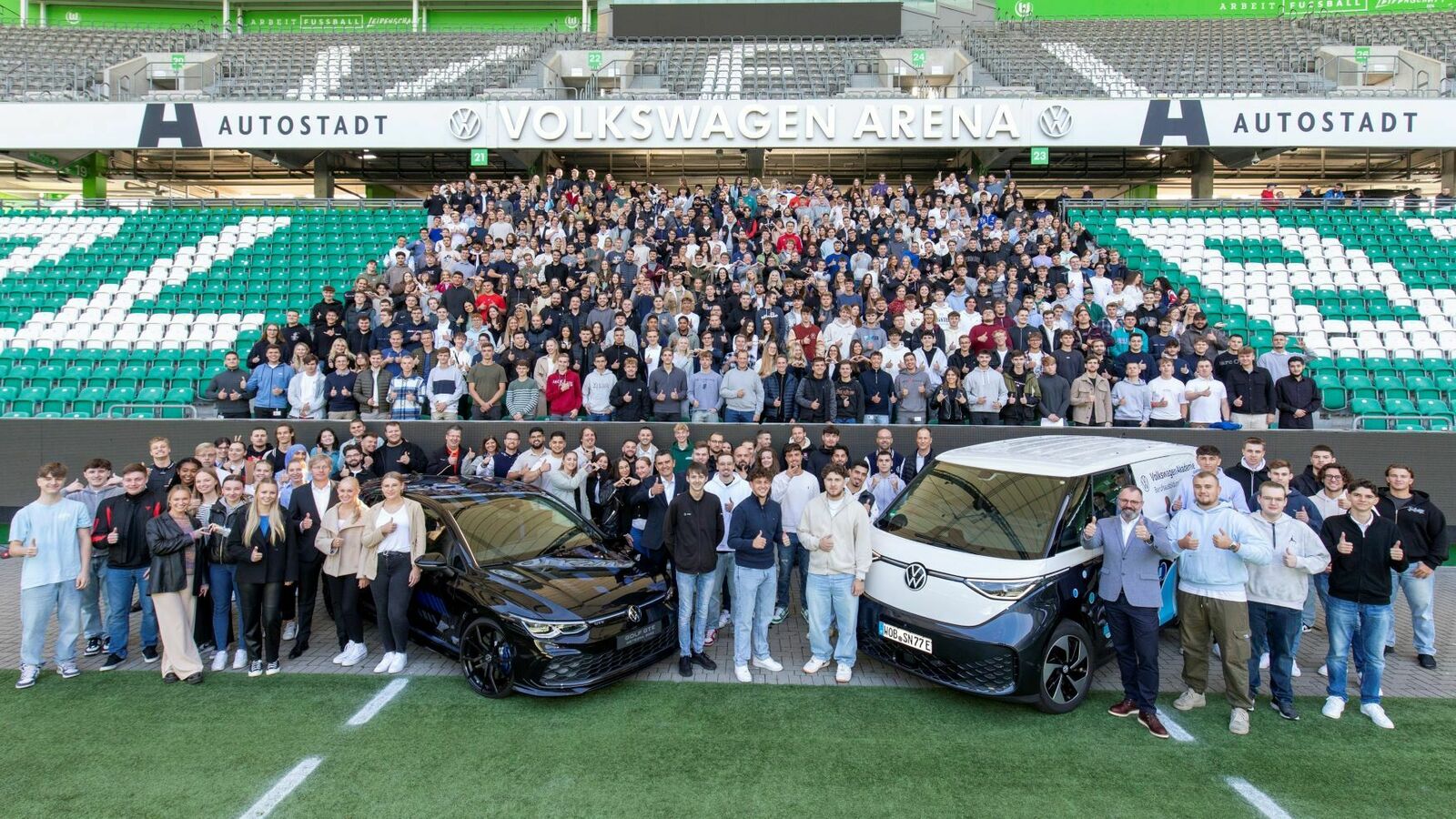 Eine große Gruppe von Menschen versammelt sich im Volkswagen Arena-Stadion für ein Gruppenfoto. Im Vordergrund stehen zwei Volkswagen-Fahrzeuge: ein schwarzer Sportwagen und ein weißer ID. Buzz mit blauem Design. Die Teilnehmer, darunter viele junge Menschen, tragen überwiegend legere Kleidung und zeigen Daumen-hoch-Gesten. Im Hintergrund sind die grünen Tribünen und der Schriftzug "Volkswagen Arena" zu sehen.
