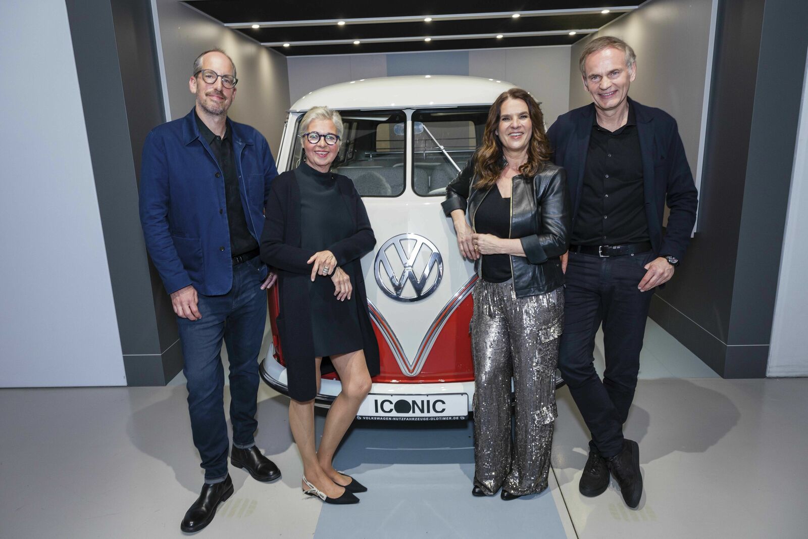Four people stand smiling next to a classic Volkswagen bus in a showroom under the "ICONIC" theme.