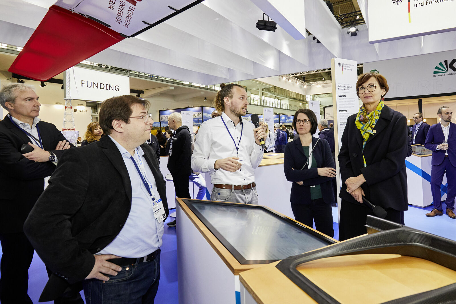 Ein Gruppe von Personen, die auf einer Messe oder Konferenz stehen, hört einem Mann zu, der ein Mikrofon hält und offenbar etwas präsentiert.