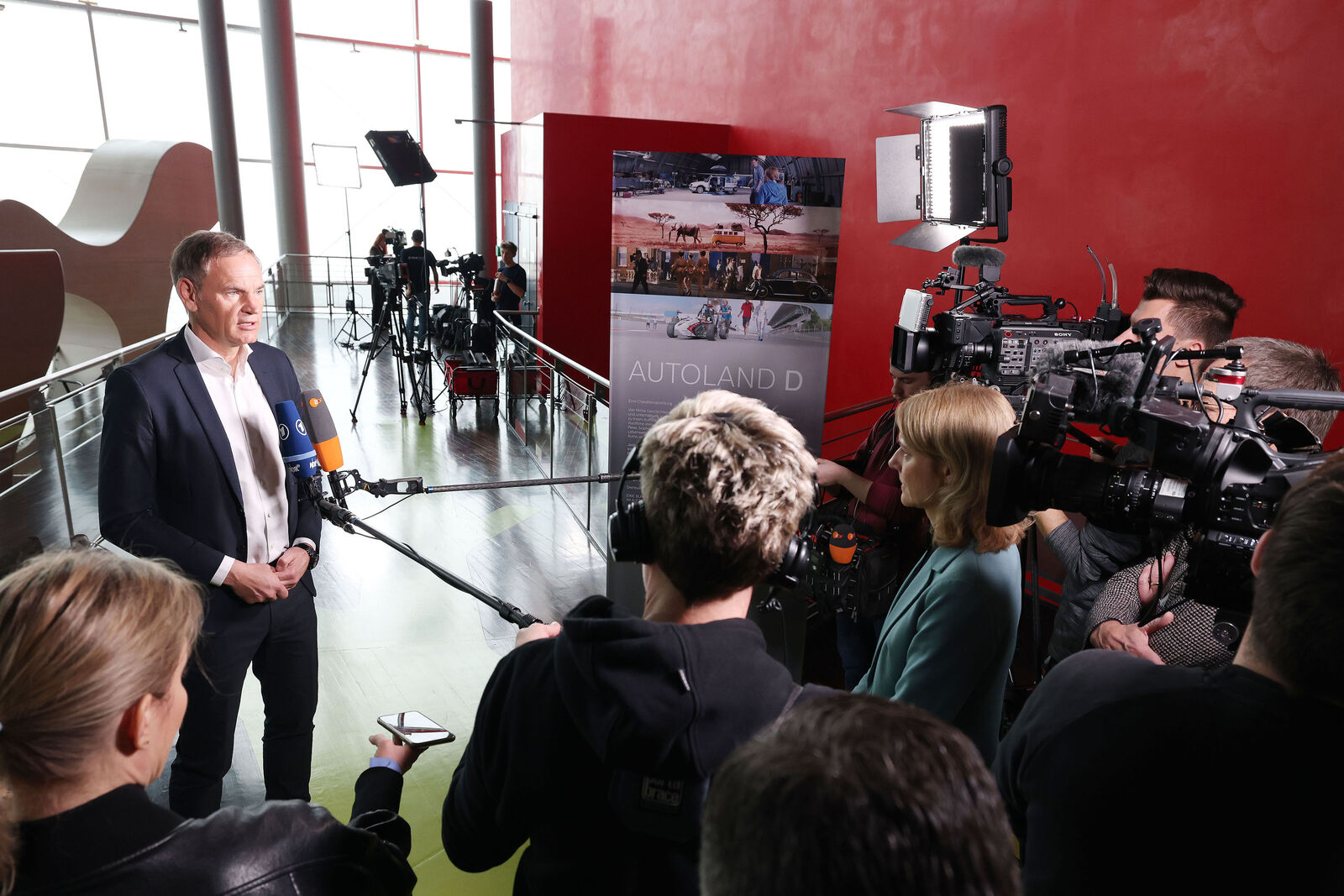 Ein Mann im Anzug gibt ein Interview vor einer Gruppe von Journalistinnen und Kameraleuten. Mikrofone, Kameras und Beleuchtung sind auf ihn gerichtet. Im Hintergrund steht ein Plakat mit der Aufschrift „AUTOLAND D“ vor einer roten Wand.